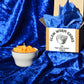 Mac and Cheez candle in a porcelain bowl with crushed velvet blue backdrop. Display also has the box that is stuffed with blue tissue paper.