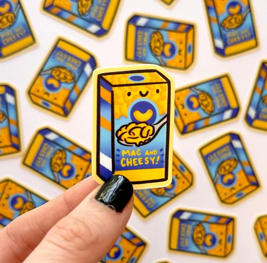 A hand with black nail polish holds a die-cut sticker shaped like a kraft mac and cheese box, with the text 'MAC AND CHEESY!' and a yellow spoonful of mac and cheese. Multiple identical stickers are scattered in the background on a white surface.