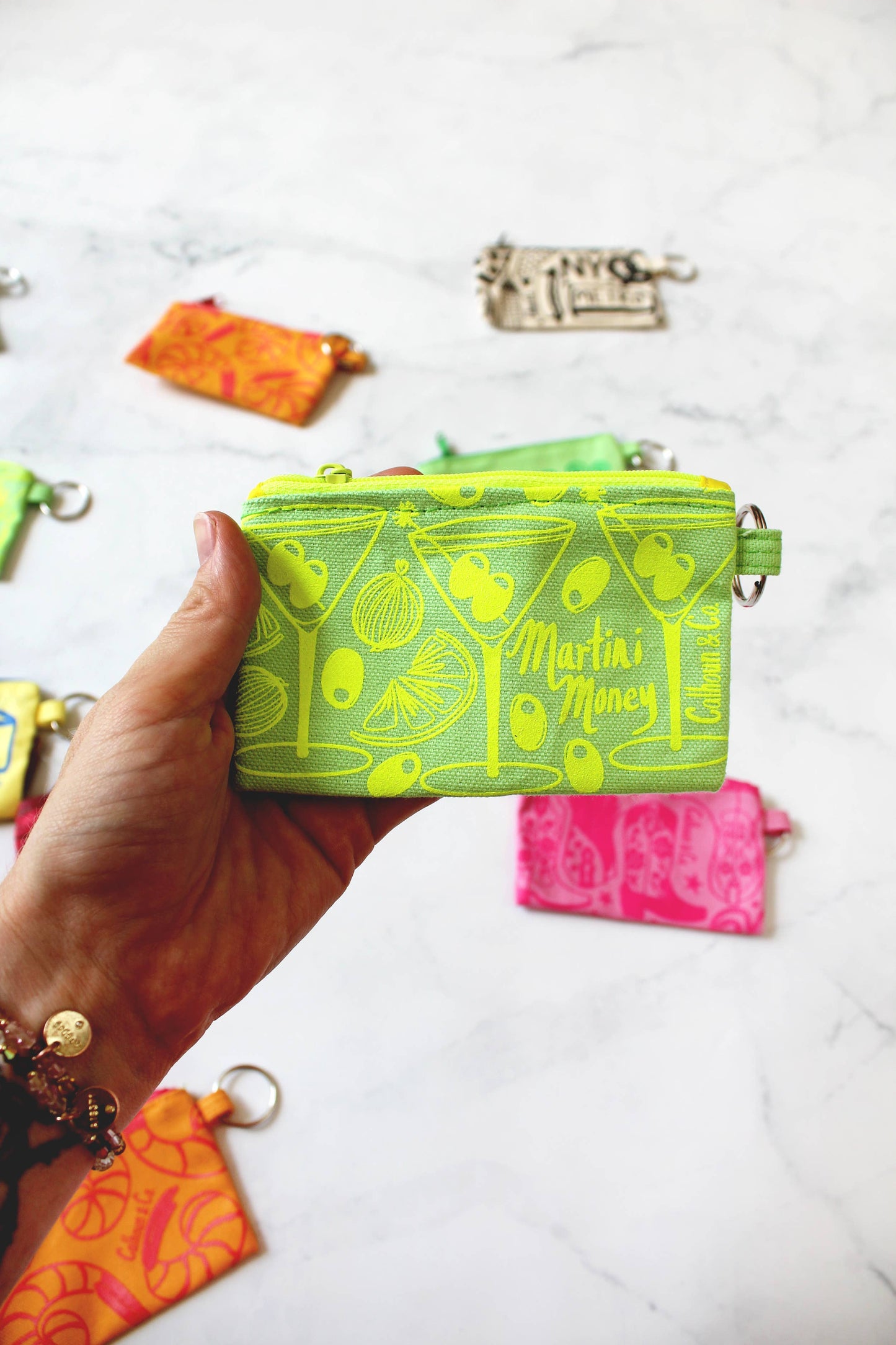 Bright green with bright lime green printing. Martini money pouches with olives, onions, and citrus. Keyring attached. text that reads: Calhoun & Co. Hand holding the bag with other pouches in the background.