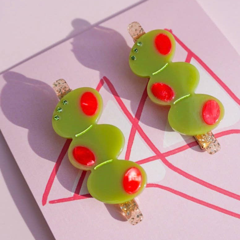 Pair of green stuffed olive hair clips with red accents on a pink background