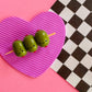 Three olives on a toothpick with faces on a pink heart-shaped cutting board against a checkered background.