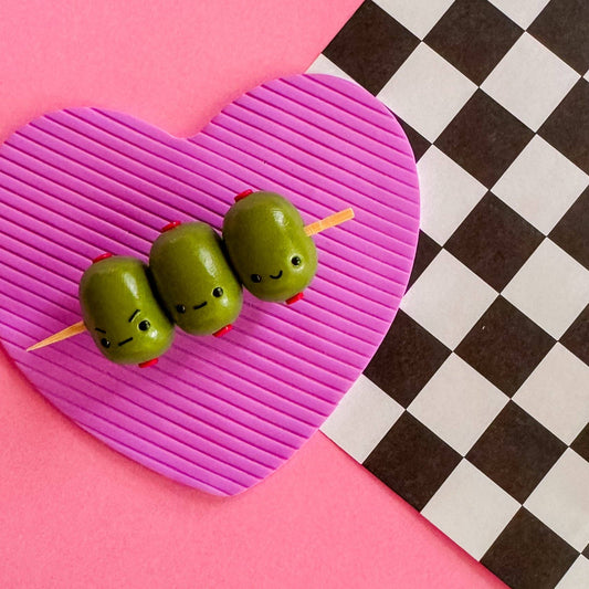 Three olives on a toothpick with faces on a pink heart-shaped cutting board against a checkered background.
