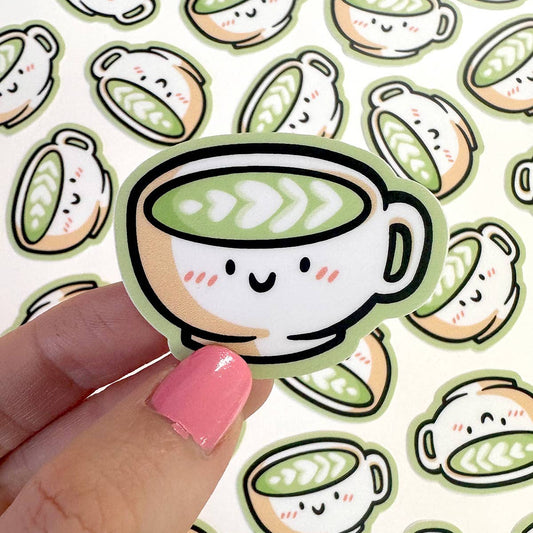 A cute die-cut sticker of a smiling matcha coffee cup is held by a hand with pink nail polish against a background of matching sticker sheets.