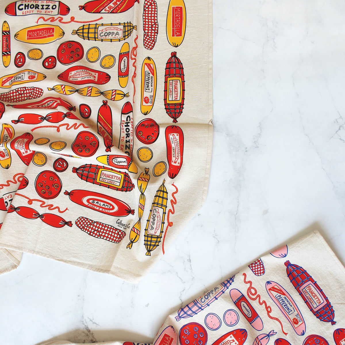 Two Meat Shop towels displaying the different color schemes. BOth towels have assorted meat, salami and cold cuts.