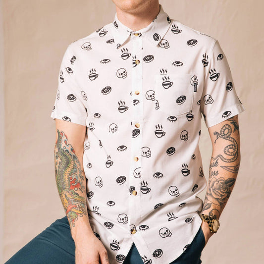 White button up shirt with tortoise buttons. Patterned with skulls, donuts, coffee and eyes. Male model with lots of tattoos.