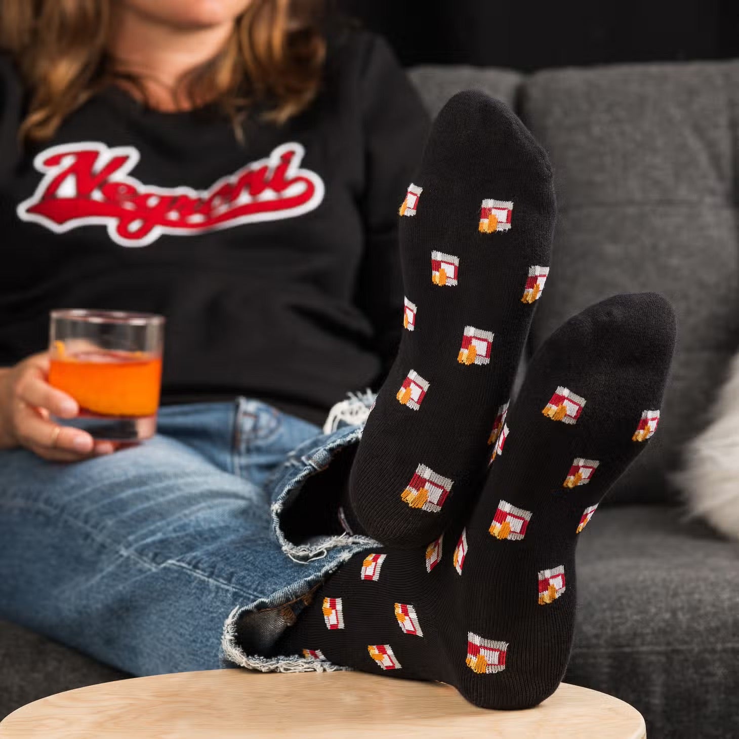 Person sitting on a couch, drinking a negroni, wearing a negroni shirt, with their feet on a coffee table to display their Negroni Socks.