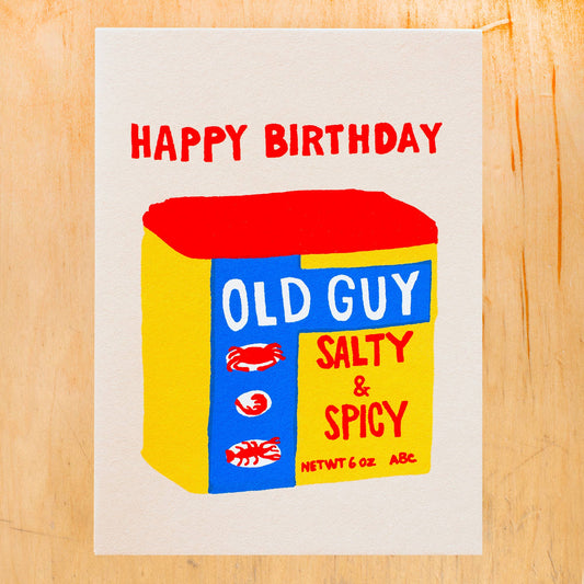 Birthday card with a box design labeled 'Old Guy Salty & Spicy' on a wooden surface.