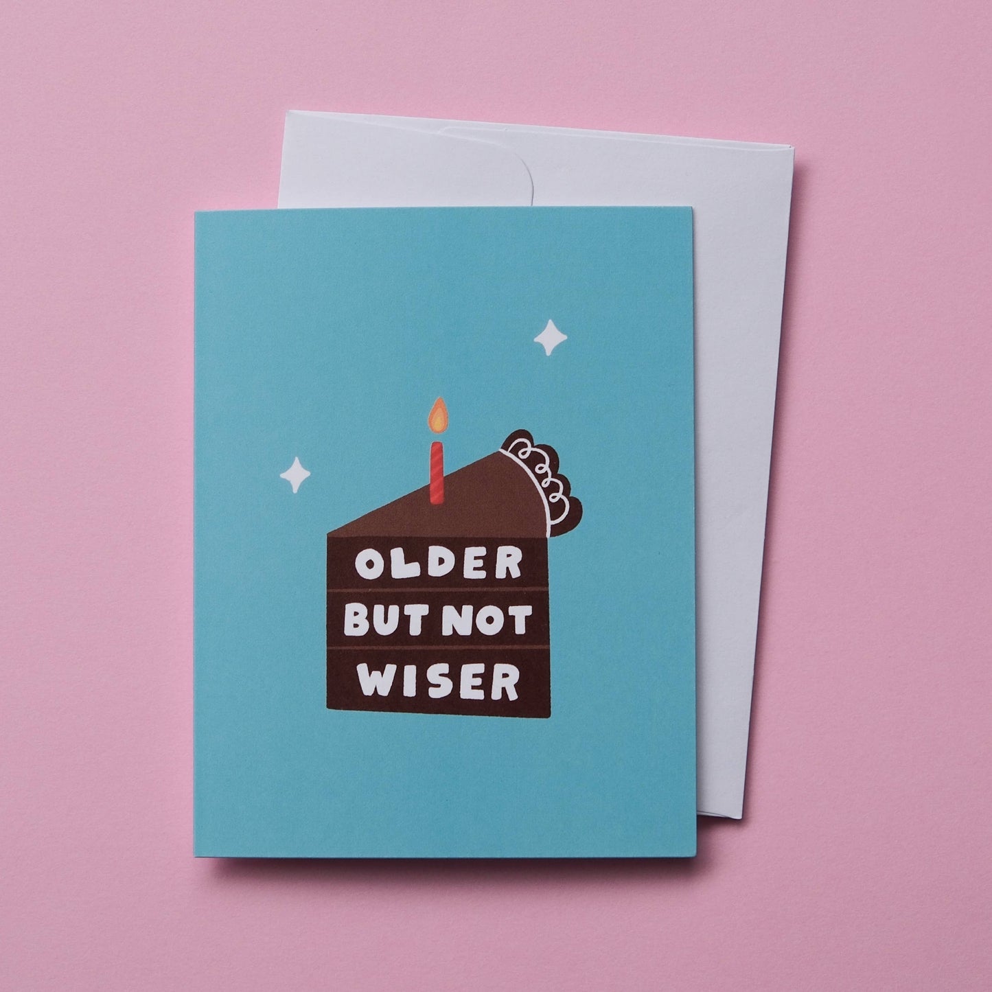 A light blue birthday card with a cartoon cake illustration and the text 'OLDER BUT NOT WISER' in white, shown with a white envelope on a solid pink background.