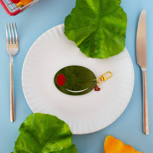 Keychain shaped like a large green olive with red filling. Gold hardware/clasp. Shown on a white plate with fork and knife on a blue tablecloth.