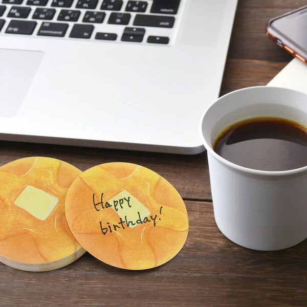 Memo pad that looks like a pancake with a melted pad of butter and syrup on it. Staged with a computer and cup of coffee with writing on the notepad that says: Happy Birthday!