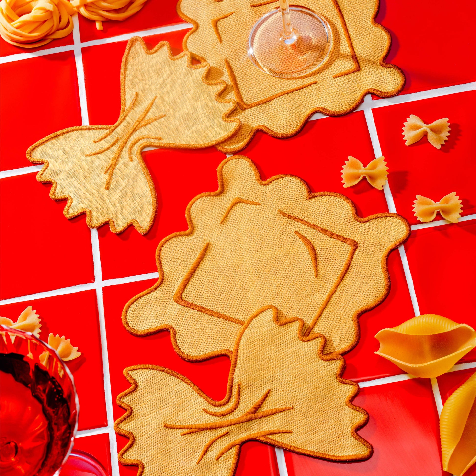 Linen pasta-shaped coasters on a red tiled surface with glasses of red wine.