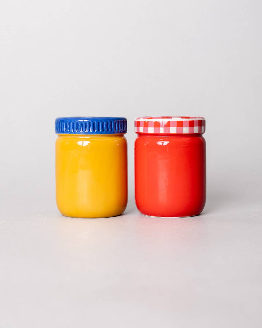 Two jars, one yellow with a blue lid and one red with a red and white checkered lid, on a light gray background.
