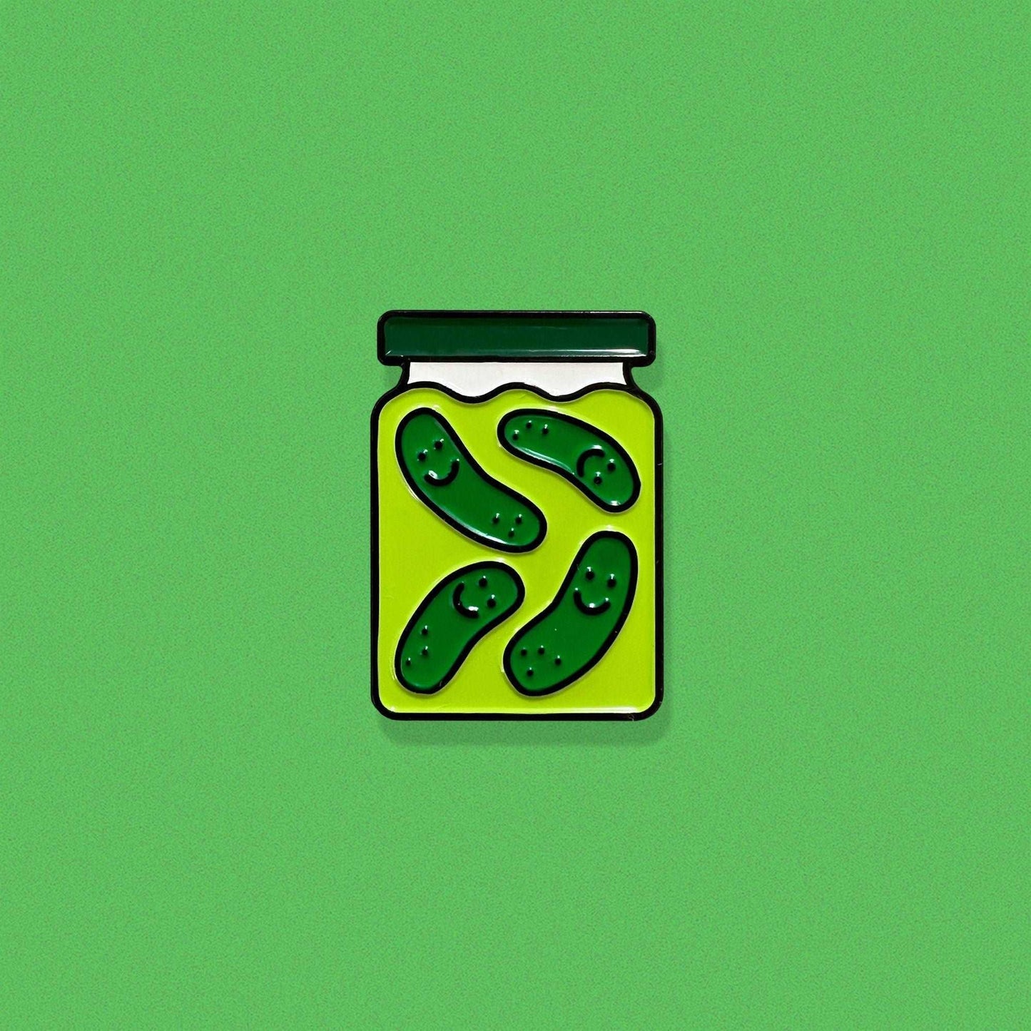 Enamel pin shaped like a pickle jar with a green lid and four smiling dill pickles floating in brine.