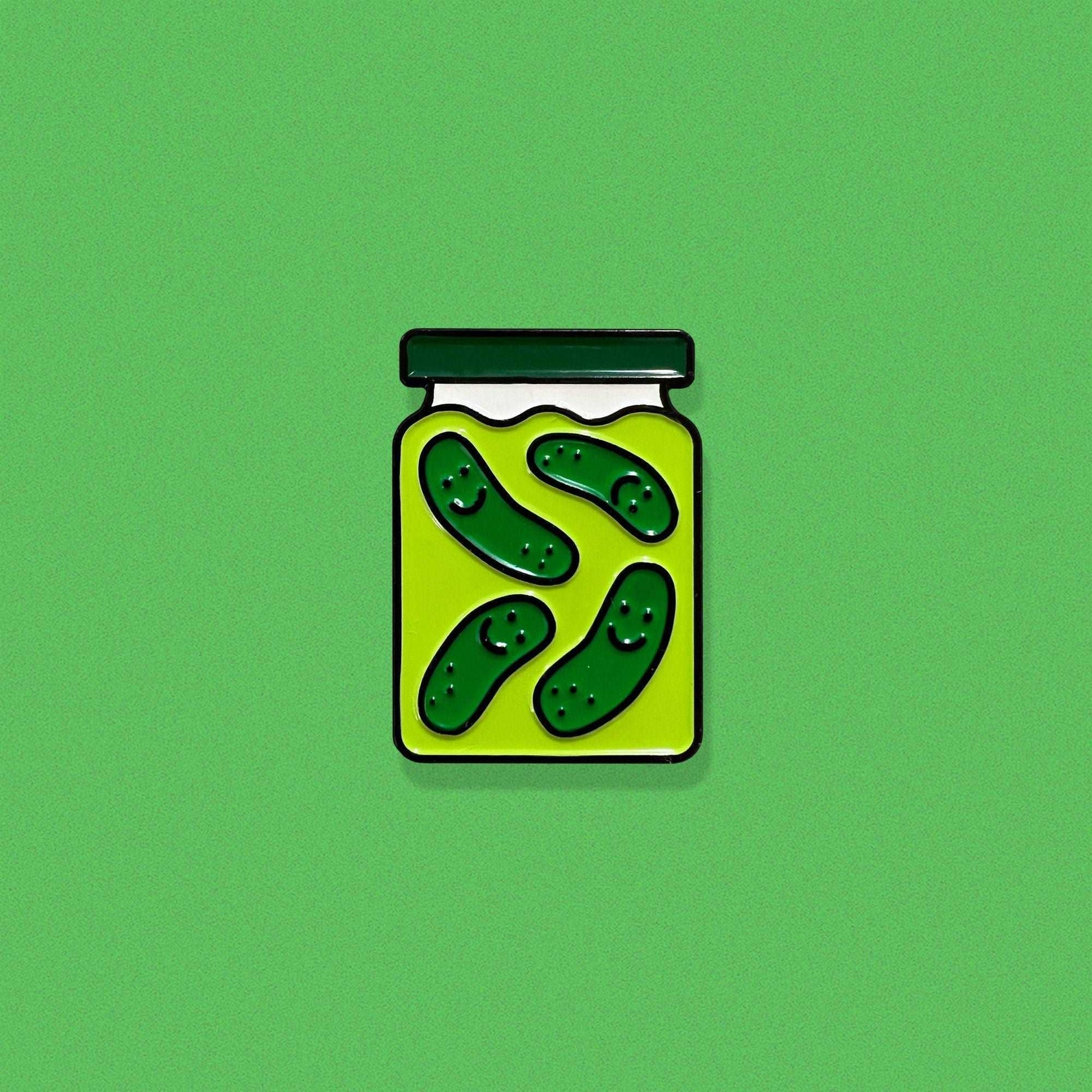 Enamel pin shaped like a pickle jar with a green lid and four smiling dill pickles floating in brine.