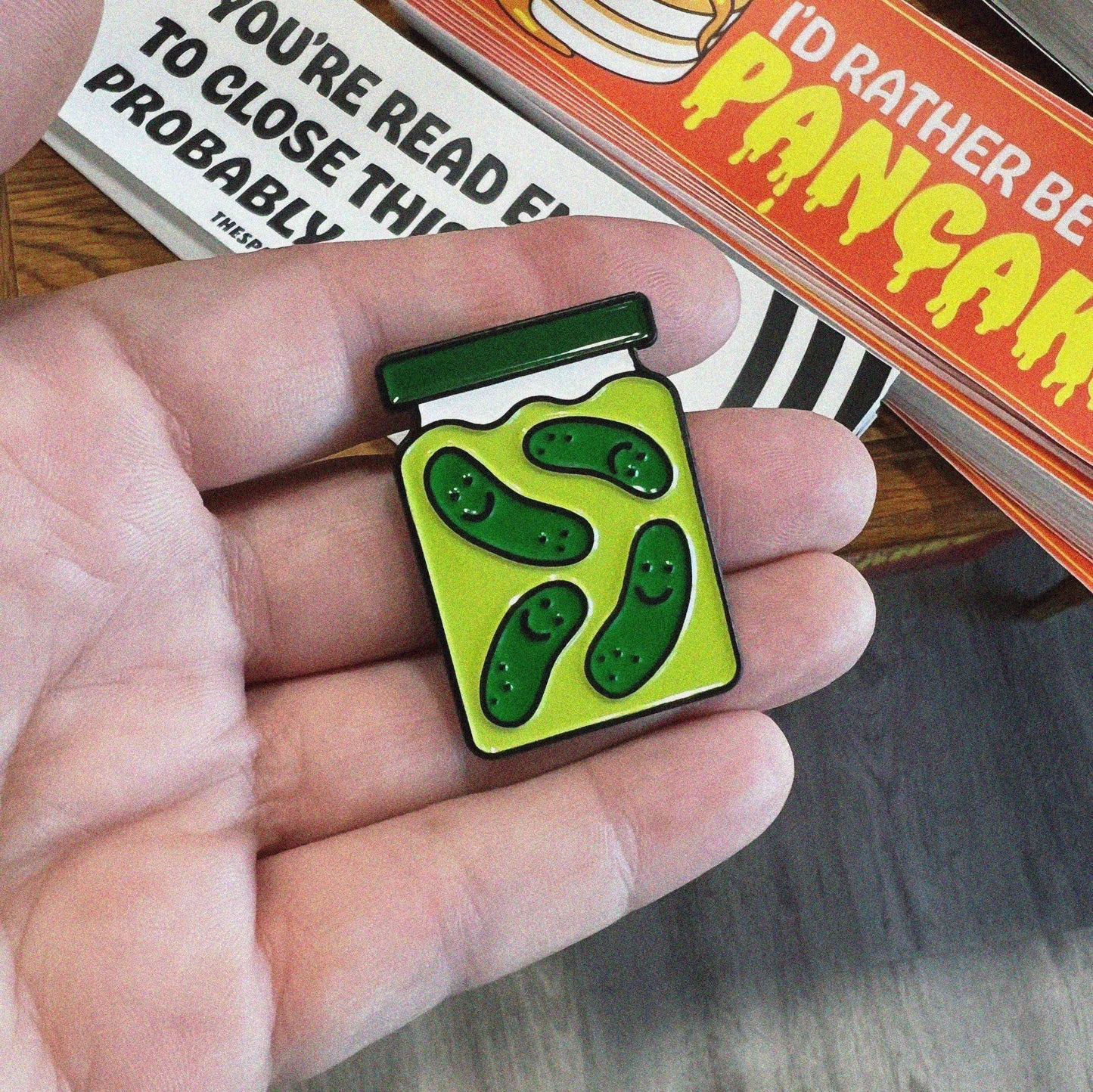 Enamel pin shaped like a pickle jar with a green lid and four smiling dill pickles floating in brine. Shown in an open hand for size comparison.