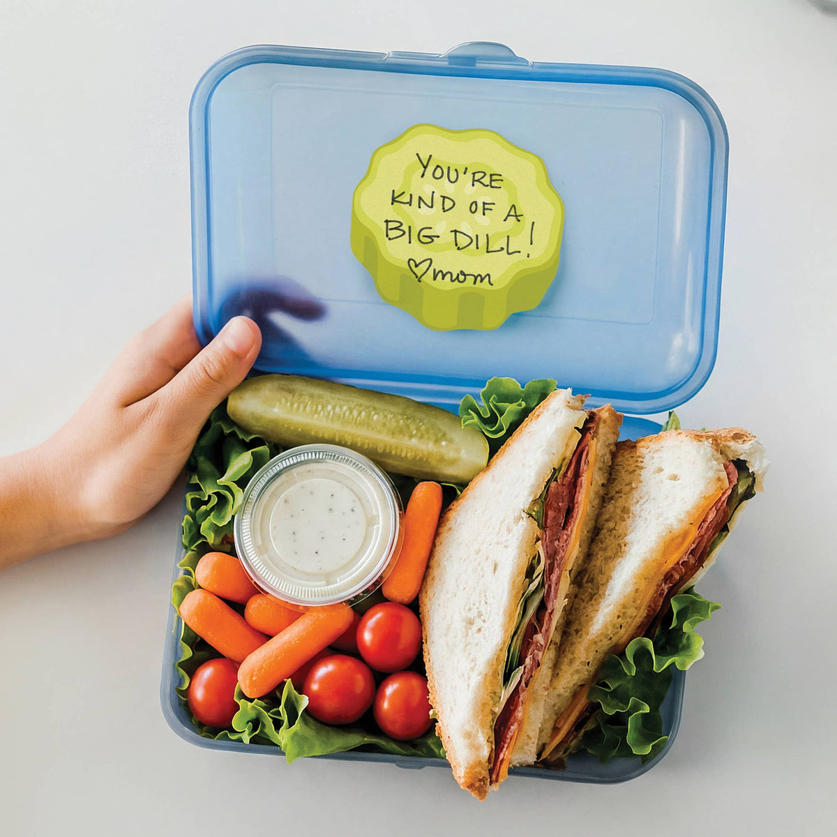 An open blue lunchbox held by a hand, filled with a sandwich, pickle, carrots, tomatoes, and a small cup of dressing. The lid has a yellow note that reads, 'You're kind of a big dill! mom'.
