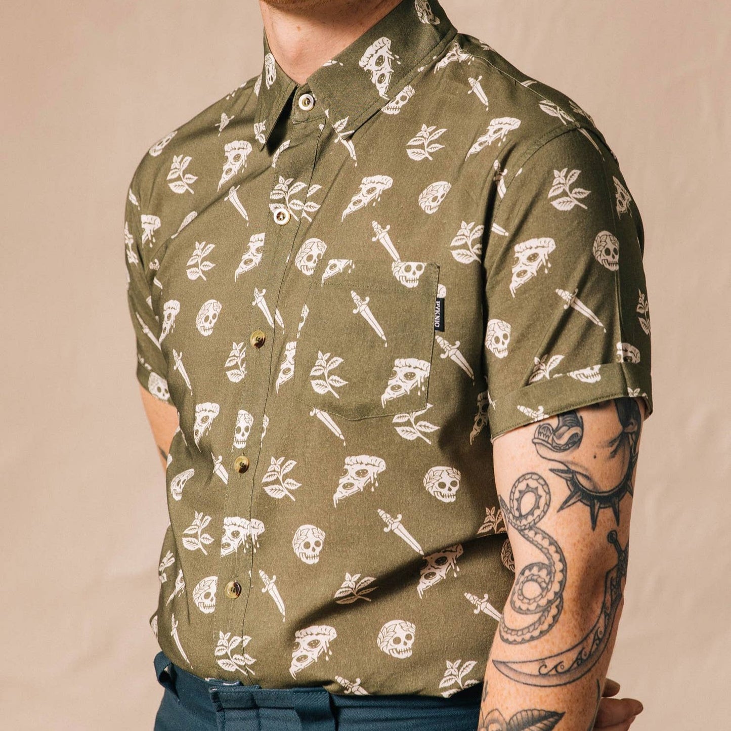 Man wearing a button up shirt that is oregano green with skulls, pizza and swords on it. 