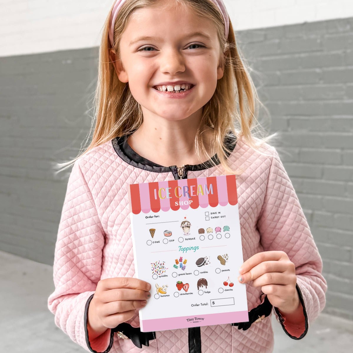 Young girl holding an ice cream shop ordering sheet against a gray wall.