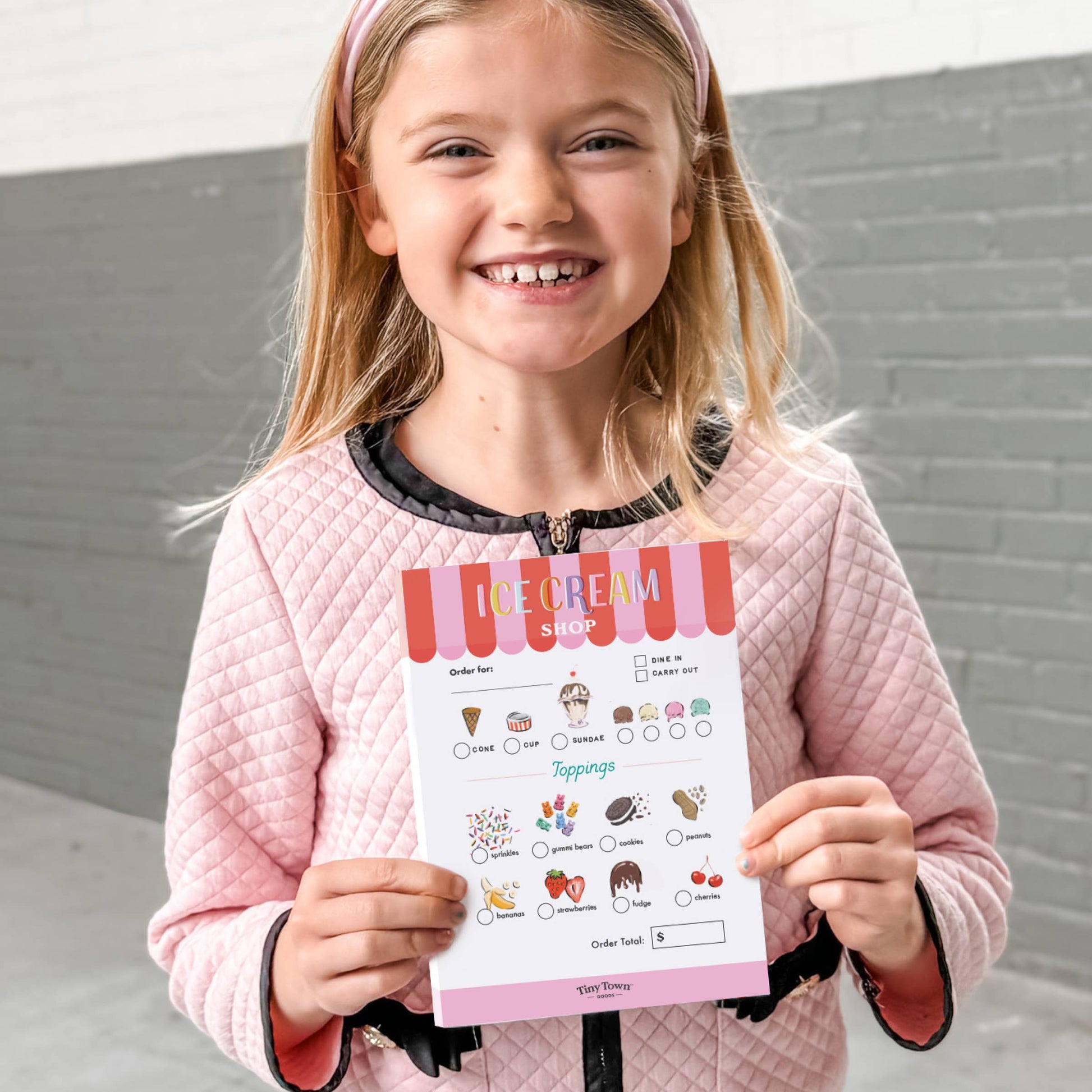 Young girl holding an ice cream shop ordering sheet against a gray wall.