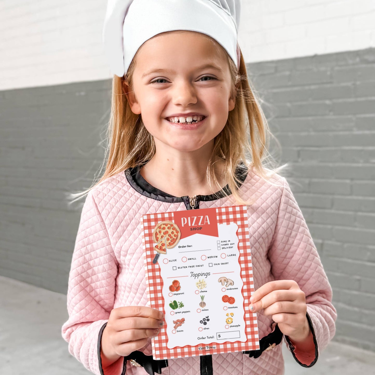 Child wearing a chef's hat and holding a pizza-making chart against a gray wall.
