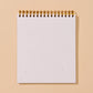 A blank white spiral-bound notepad with a gold wire binding, standing upright against a solid warm beige background.