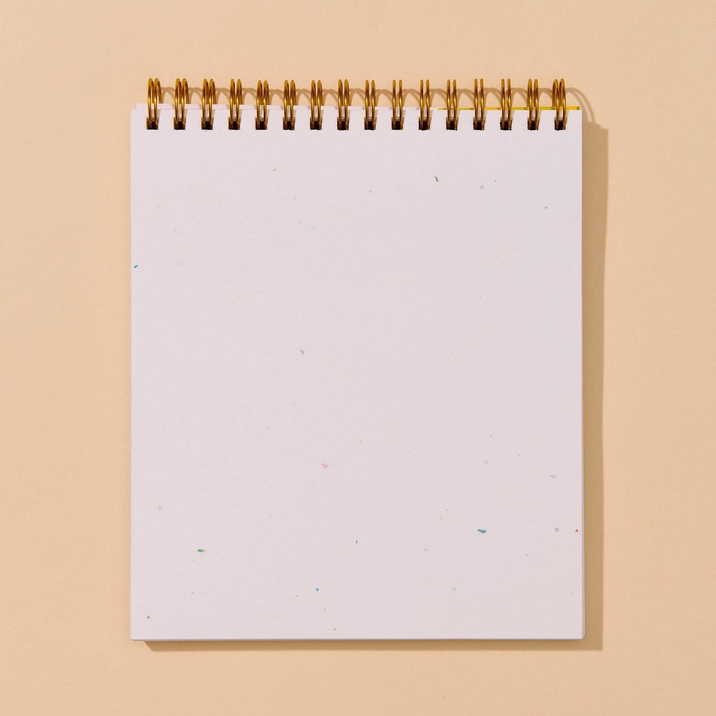 A blank white spiral-bound notepad with a gold wire binding, standing upright against a solid warm beige background.