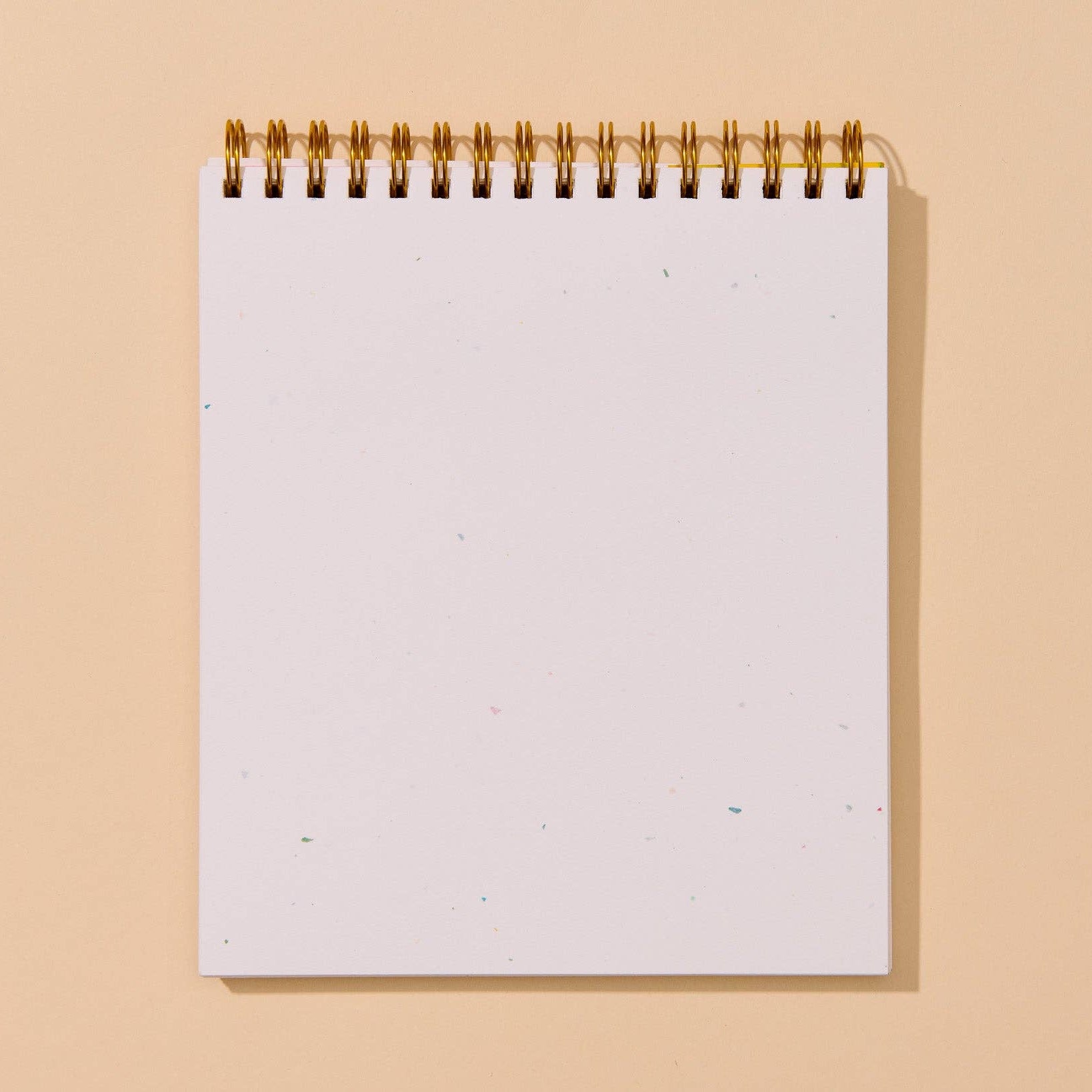 A blank white spiral-bound notepad with a gold wire binding, standing upright against a solid warm beige background.