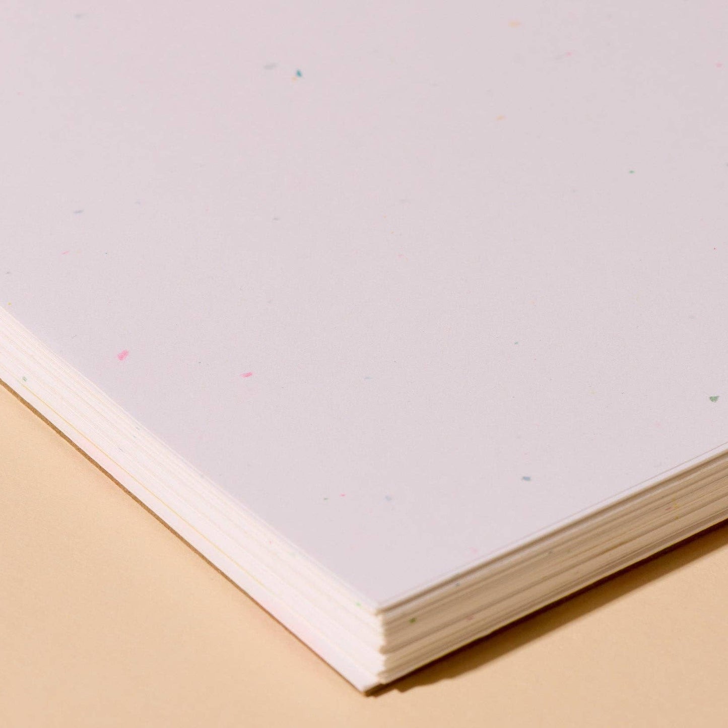 A close-up of a stack of white paper sheets with tiny multicolored flecks, resting on a warm beige surface.