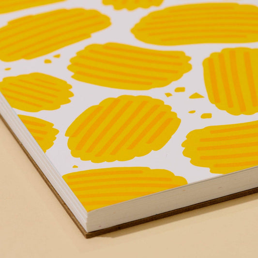 A close-up of a notebook or book page with a white cover patterned with bright yellow oval potato chip shapes, photographed at an angle on a light beige surface.