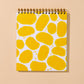 A spiral-bound notebook with a white cover decorated with a pattern of yellow potato chips, displayed against a solid beige background.