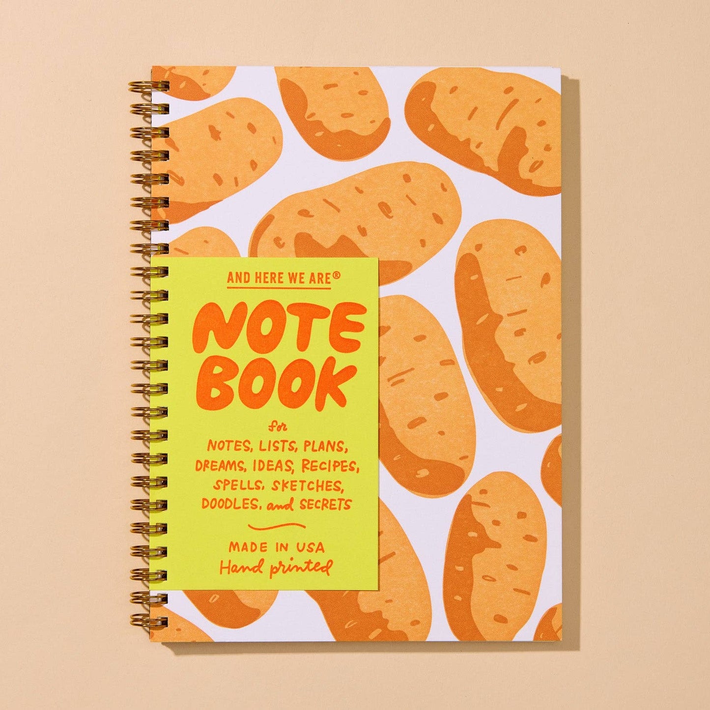 A spiral-bound notebook with a white cover decorated with a pattern of orange-brown potatoes, featuring a bright yellow front cover with the text 'AND HERE WE ARE! NOTE BOOK'.