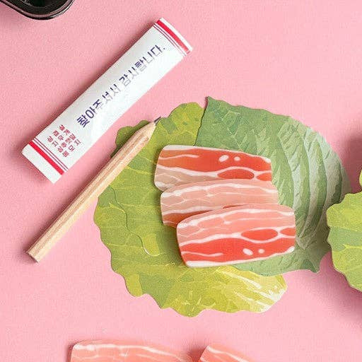 Erasers that look like slices of raw meat on green leaf-shaped memo note with a pencil and tube on a pink background