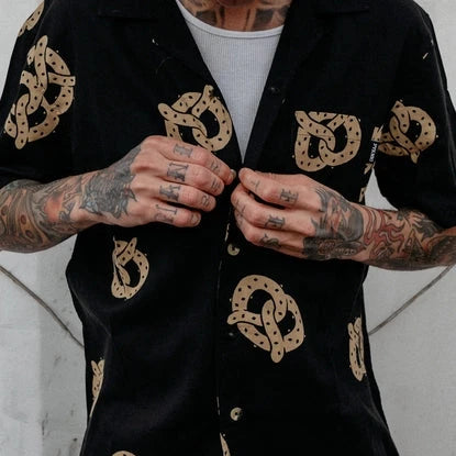 Person with heavily tattooed arms buttoning up a black shirt with all-over pretzels print.