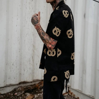 Photo of man with tattoos on his arm wearing button up pretzel shirt and black jeans.