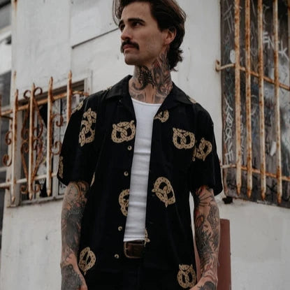 Photo of man with tattoos on his arms wearing pretzel shirt, unbuttoned over a white undershirt and black jeans.