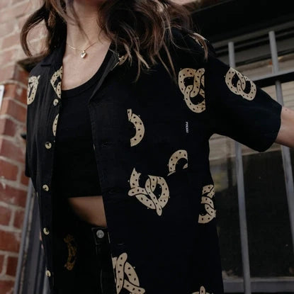 Photo of woman with long hair wearing pretzel shirt unbuttoned over black tank top, with black jeans.