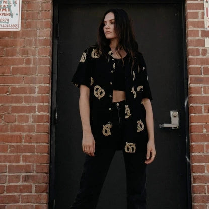 Photo of woman with long hair wearing pretzel shirt unbuttoned over black crop top, with black jeans and sneakers.