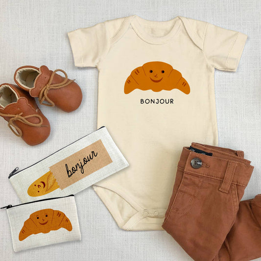 flat lay of the bonjour croissant baby onesie, brown baby pants and shoes and two small pouches one with a baguette and the other with the same smiley croissant on it 