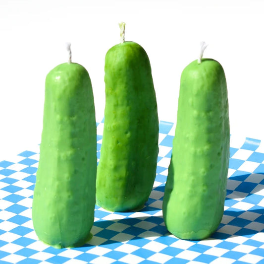 3 dill pickle candle standing up on one end. On top of blue and white checkered paper. 