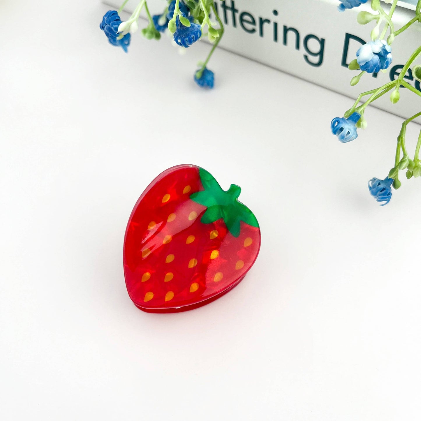 Strawberry-shaped mini hair claw on a white surface with blue flowers and text in the background