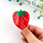 Hand holding a strawberry-shaped hair claw with a white background and blue flowers.