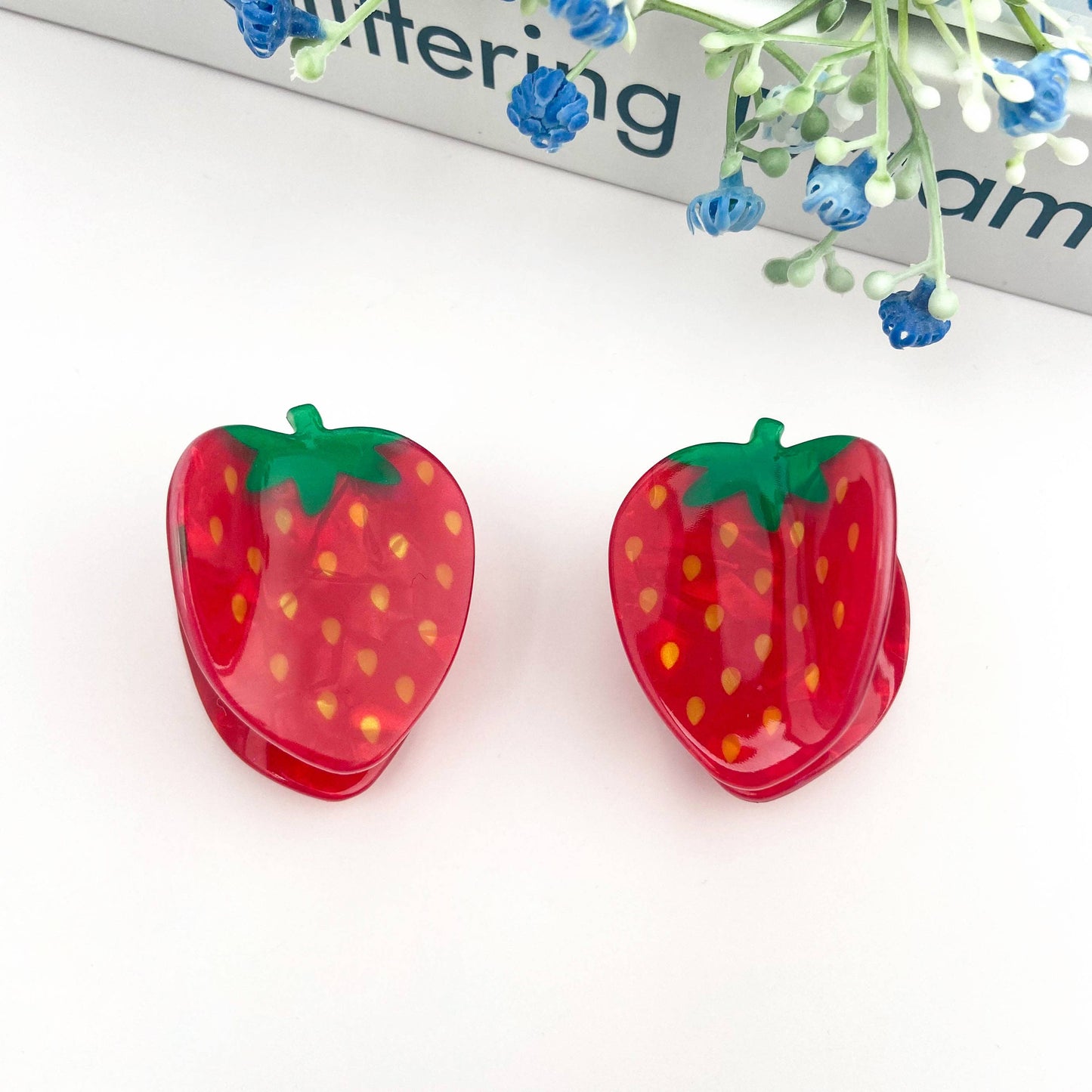 Strawberry-shaped hair clips on a white background with a magazine in the background.