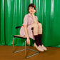 A woman sits on a metal chair in front of a bright green curtain backdrop. She is wearing a pink and white vertically striped short-sleeve collared dress with a red waist tie, dark patterned knee-high socks, and light pink slip-on shoes.