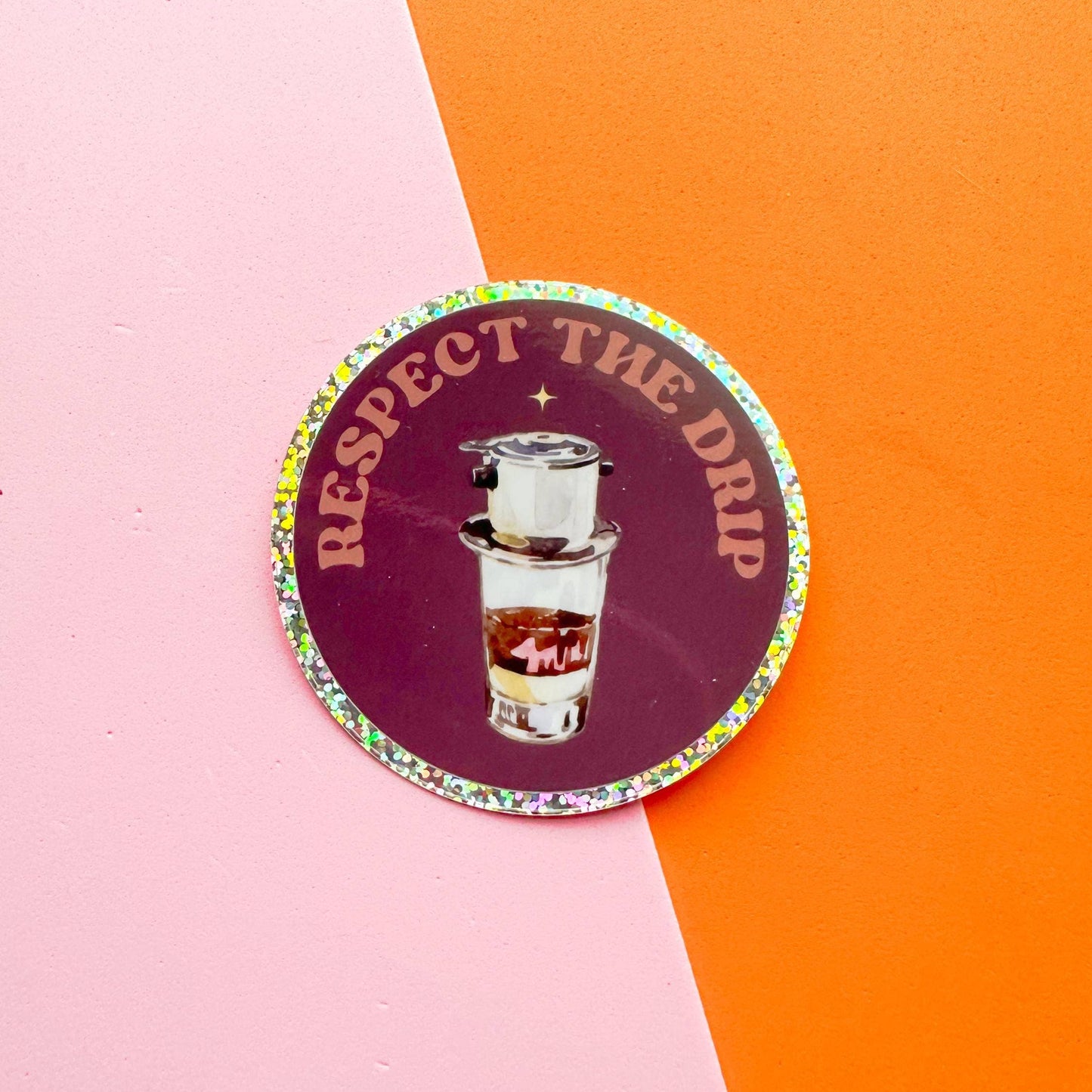 A round sticker with a glittery border featuring the text 'RESPECT THE DRIP' in gold around a central illustration of a coffee pour-over setup, placed on a diagonally split pink and orange background.