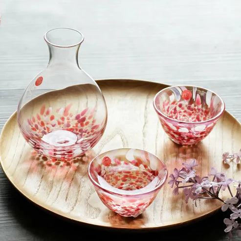 Set of glass sake carafe and two cups with speckled red and white patterns on a gold tray