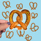 A hand with purple nail polish holds a die-cut sticker shaped like a pretzel, with the words 'Salty' and 'Bitch' written in yellow "mustard" across it. The background is light blue and decorated with multiple smaller pretzel outline stickers.