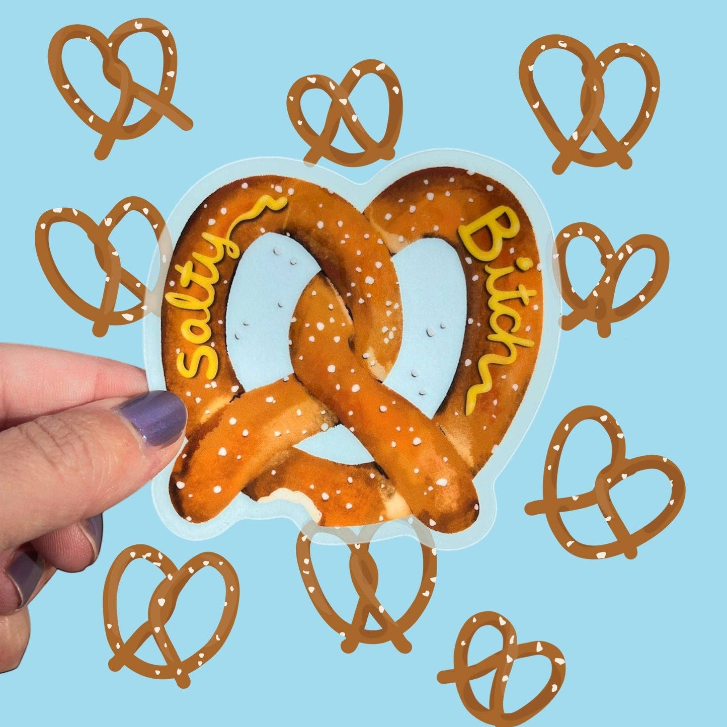 A hand with purple nail polish holds a die-cut sticker shaped like a pretzel, with the words 'Salty' and 'Bitch' written in yellow "mustard" across it. The background is light blue and decorated with multiple smaller pretzel outline stickers.
