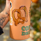 A hand holds a tall insulated tumbler with a peach-orange finish. The tumbler features a die-cut sticker of a pretzel with the words 'Salty Bitch' and green text reading 'COFFEE DRINK'.
