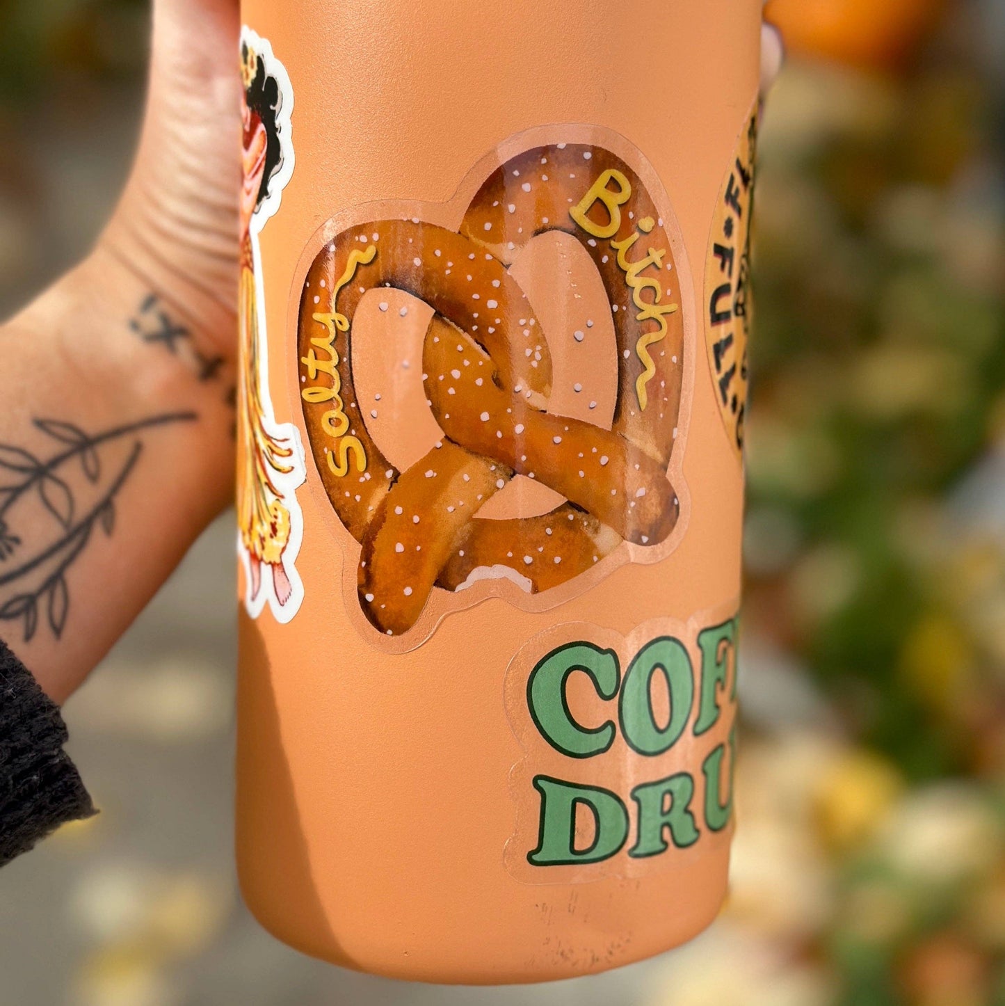 A hand holds a tall insulated tumbler with a peach-orange finish. The tumbler features a die-cut sticker of a pretzel with the words 'Salty Bitch' and green text reading 'COFFEE DRINK'.