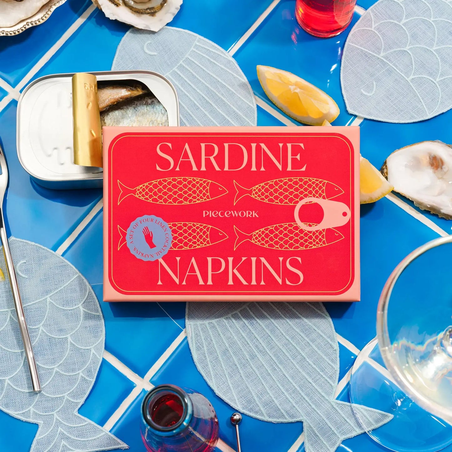 Sardine napkin box set on a table with blue tile, sardines in a bin, lemon wedges and napkins scattered.